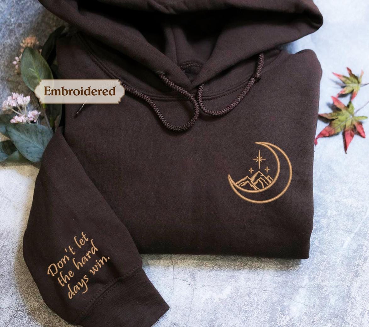 A brown hoodie with a crescent moon and stars embroidered on it, along with a small text message on the sleeve.