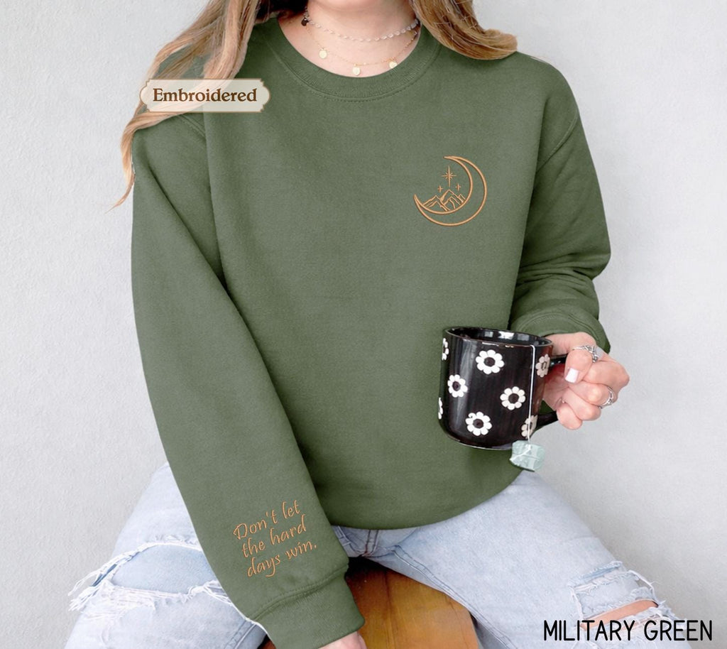 A woman wearing a green sweater with a crescent moon and star design, holding a black mug with white flowers, and a text embroidered on the sleeve of the sweater.