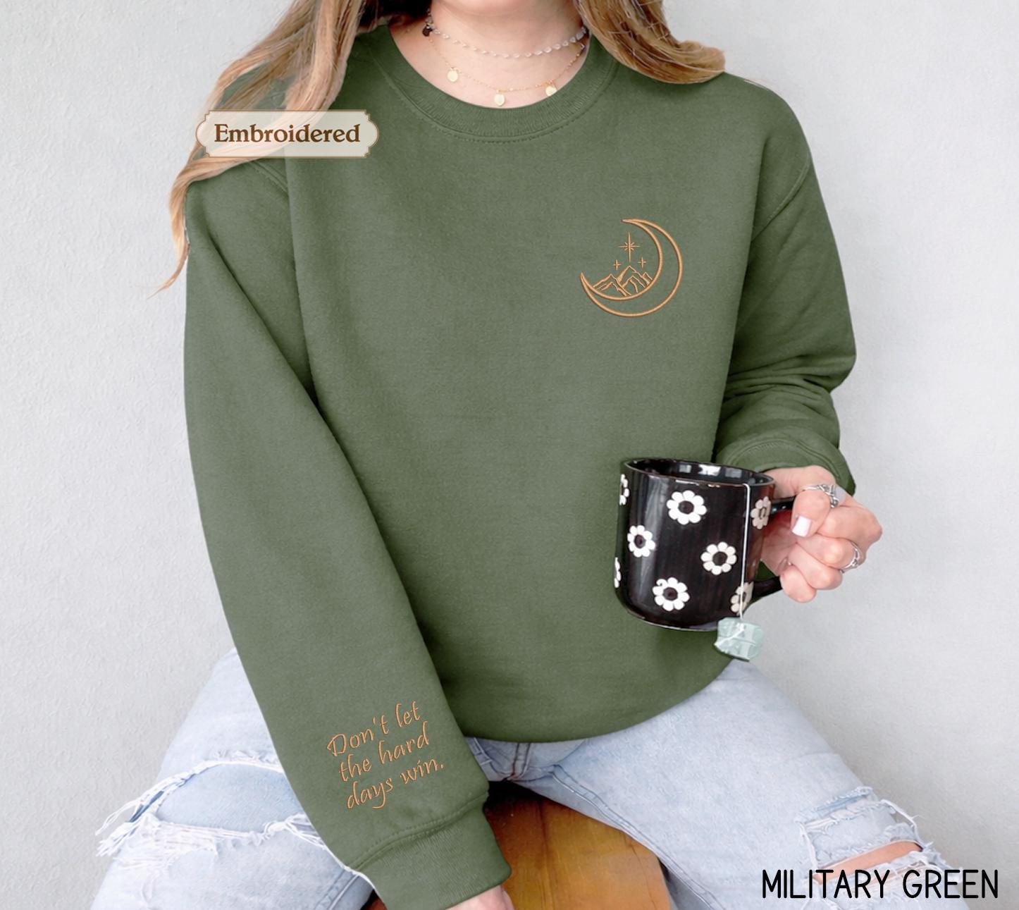 A woman wearing a green sweater with a crescent moon and star design, holding a black mug with white flowers, and a text embroidered on the sleeve of the sweater.