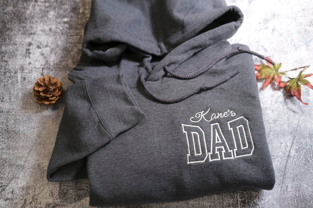 A gray hoodie with the text "Kane's DAD" printed on it, along with a pine cone and some leaves, is displayed on a textured surface.