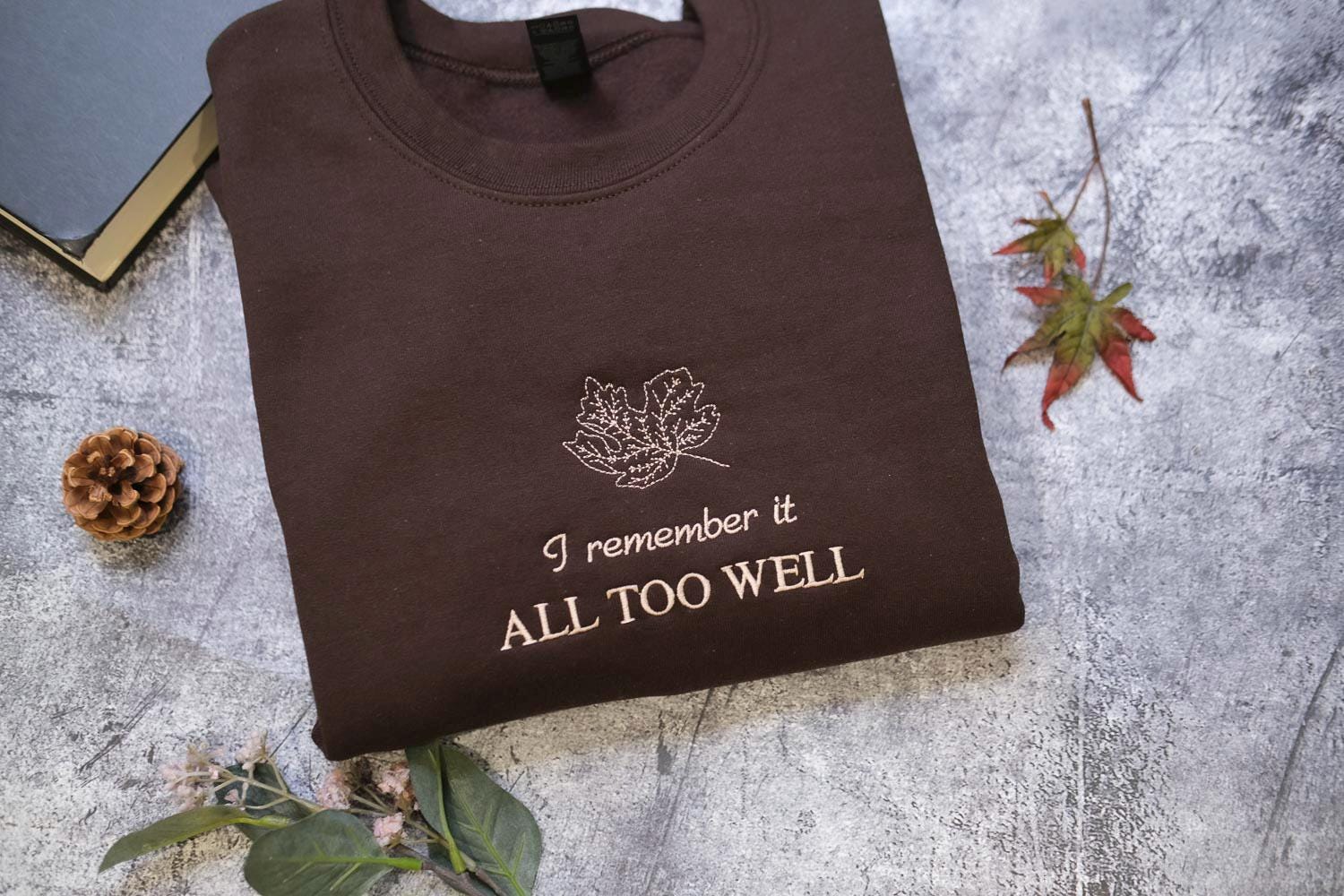 a brown sweatshirt with a leaf design and the text "I remember it all too well" printed on it, along with a pine cone and a small leaf on a gray surface.