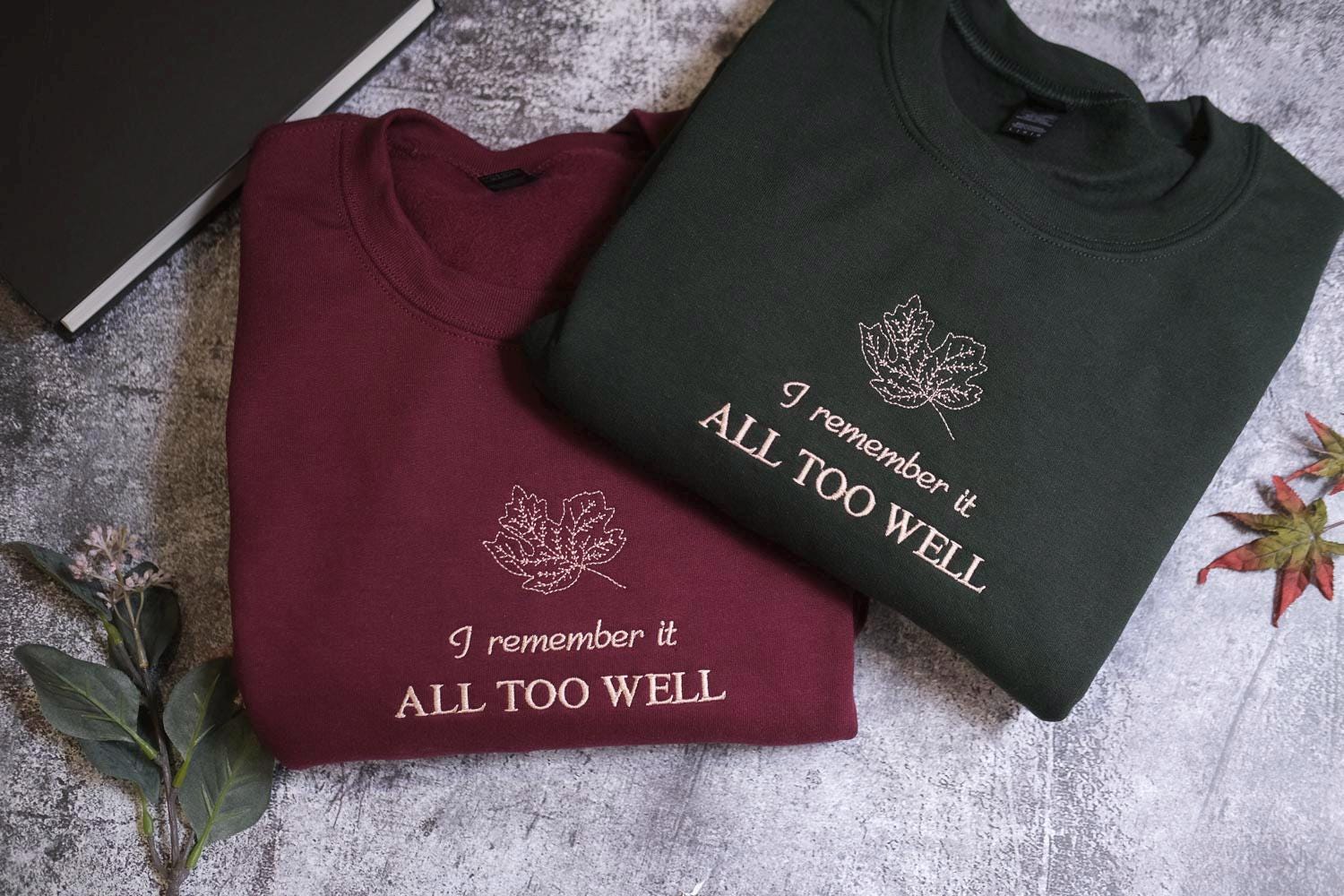 two t-shirts with a leaf design, one burgundy and one dark green, placed on a gray surface.