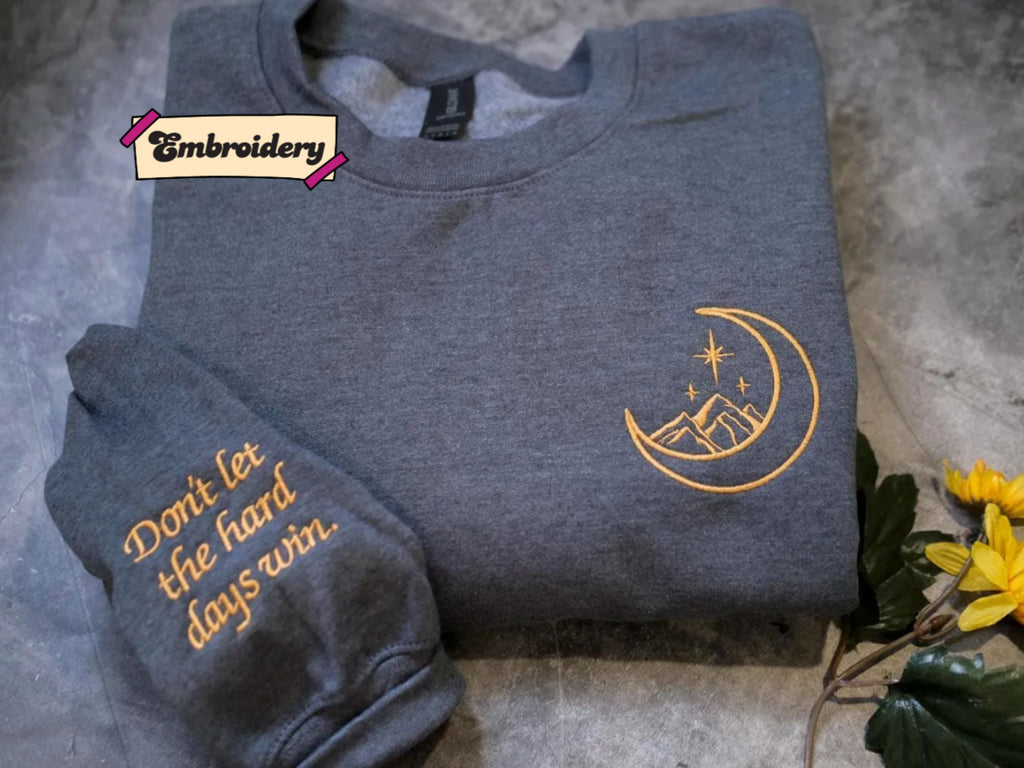 a gray sweatshirt with an embroidered design on the right side, featuring a crescent moon and stars. The sweatshirt is placed on a gray surface, and there is a small yellow flower to the right of the sweatshirt.