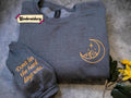 a gray sweatshirt with an embroidered design on the right side, featuring a crescent moon and stars. The sweatshirt is placed on a gray surface, and there is a small yellow flower to the right of the sweatshirt.