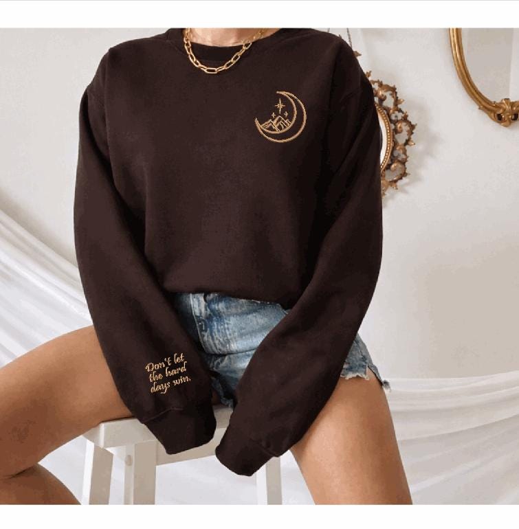A person wearing a black sweatshirt with a crescent moon design and a gold chain necklace, sitting on a white stool against a white background.