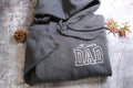 A gray hoodie with the text "Kane's DAD" printed on it, along with a pine cone and a small leaf on a textured surface.