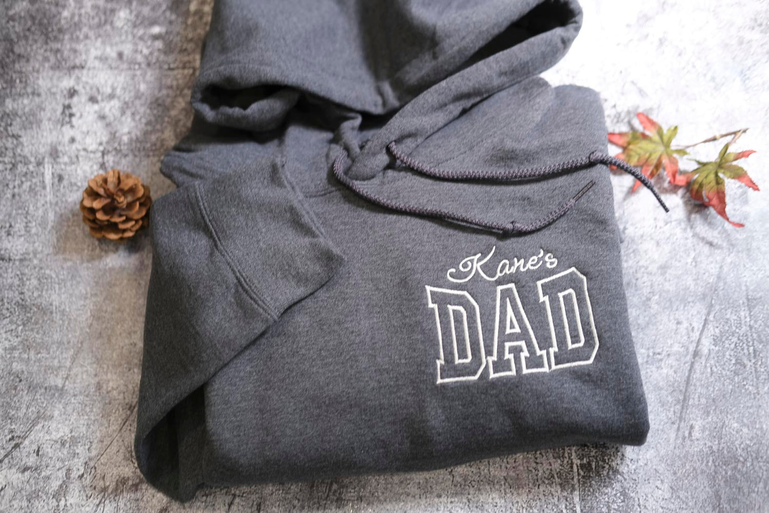 A gray hoodie with the text "Kane's DAD" printed on it, along with a pine cone and a small leaf on a textured surface.